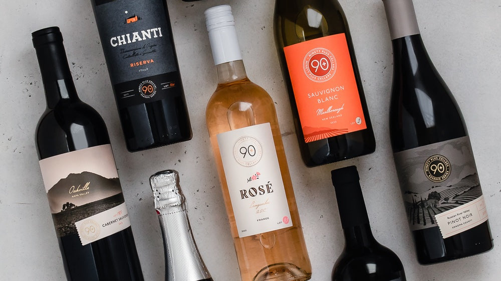 Seven mixed bottles of red and white wine on a white marble countertop. The wines shown include Lot 207 Chianti from Italy, Lot 2 Sauvignon Blanc from Marlborough, New Zealand, Lot 33 Rosé from Languedoc, France, and Lot 75 Pinot Noir from Russian River Valley, Sonoma County California.