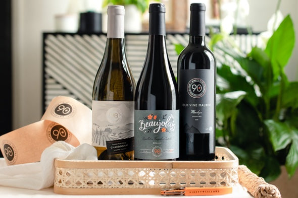 A bottle of Lot 130 Chardonnay, Lot 158 Beaujolais, and Lot 23 Malbec next  to each other in a small basket in front of a plant in the living room.